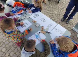 Na zdjęciu dzieci bawiące się przy policyjnym stoisku. Trzy dziewczynki i dwóch chłopców rysujących kolorowanki.