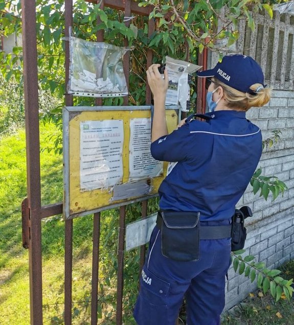 Policjantka zawieszająca ulotkę na tablicy ogłoszeń przed bramą orodów działkowych