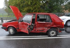 Uszkodzony samochód koloru czerwonego. Pojazd ma otwartą przednią klapę, oraz wgnieciony lewy bok.