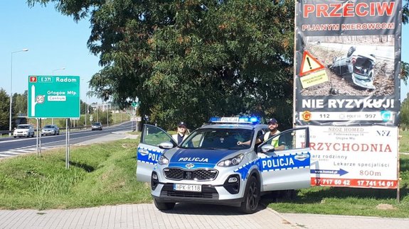 Na zdjęciu dwoje dzielnicowych z komisariatu w Głogowie Małopolskim. Policjanci stoją przy oznakowanym radiowozie na tle baneru profilaktycznego. na banerze hasło działań &amp;quot;przeciw pijanym kierowcom&amp;quot;, nie ryzykuj i zdjęcie rozbitego samochodu.