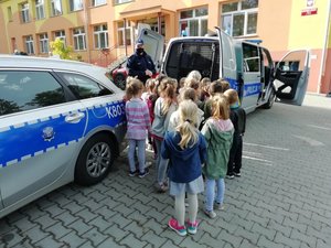 Dzieci stoją przed  dwoma radiowozami. Przed nimi policjant. W tle widoczny budynek przedszkola.