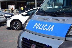 Na pierwszym planie widać przód policyjnego radiowozu, na którym jest napis Policja. Obok niego stoją trzy inne pojazdy, zaparkowane na sklepowym parkingu. W tle widać wejście do sklepu i wchodzących klientów.