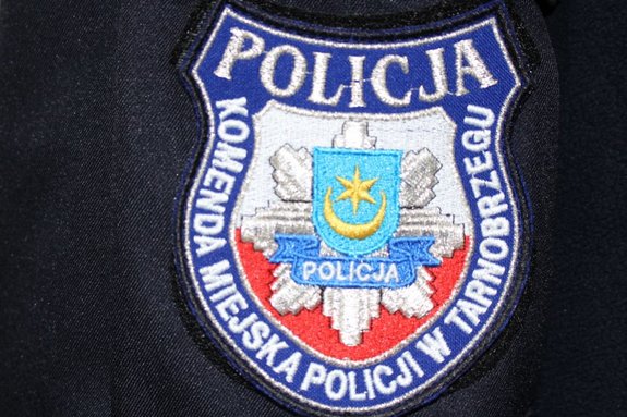 Zdjęcie przedstawia emblemat z napisem Policja - Komenda Miejska Policji w Tarnobrzegu. W środku naszywki, na biało - czerwonym tle, widnieje hebr miasta Tarnobrzega oraz napis Policja.
