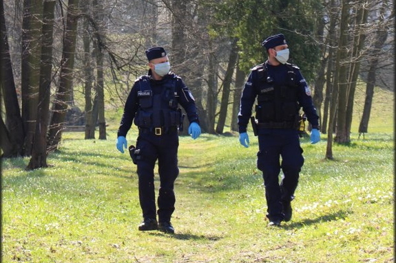Dwóch umundurowanych policjantów w maseczkach ochronnych idzie leśną ścieżką. Zdjęcie wykonane w ciągu dnia.