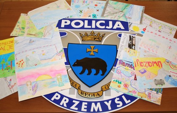 Zdjęcie kolorowe -przedstawia prace plastyczne dzieci i młodzieży które są porozkładane na emblemacie policyjnym Miasta Przemyśl. Widiczny na emblemacie jest napis (na górze ) POLICJA , poniżej jest widoczny herb miasta Przemyśl tz korona oraz niedzwiedz a poniżej zamieszczony jest napis Przemyśl