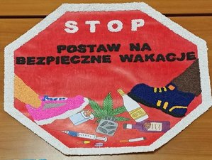 Zdjęcie kolorowe na nim obraz znaku drogowego STOP w którym namalowane są stopu przygniatające alkohol , papierosy i inne używki i napis &amp;quot;STOP POSTAW NA BEZPIECZNE WAKACJE&amp;quot;.