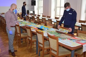 Jury konkursu wybiera najlepsze prace plastyczne - wszystkie prace rozłożone są na stołach w auli Komendy Wojewódzkiej Policji w Rzeszowie. Po prawej stronie policjant ogląda prace, w tle drugi policjant. Po lewej stronie stoi mężczyzna.