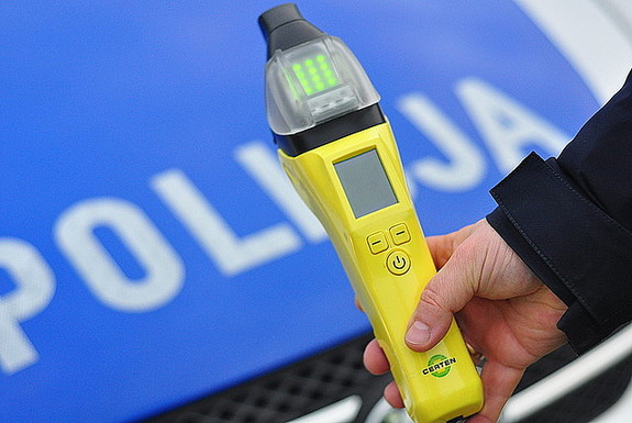 Zdjęcie kolorowe przedstawia maskę silnika radiowozy w kolorze niebieskim z napisem białym POLICJA. przed maska widoczna jest ręka prawa f-sza który trzyma alcosensor w kolorze żółtym.