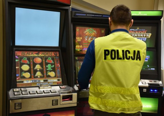 Trzy automaty do gier hazardowych. Przed nimi stoi policjant w kamizelce z napisem Policja
