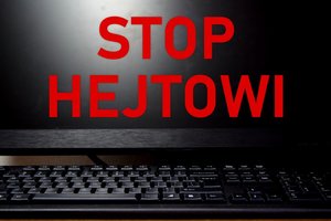 Na pierwszym planie czerwony napis Stop Hejtowi, w tle czarna klawiatura i monitor komputerowy