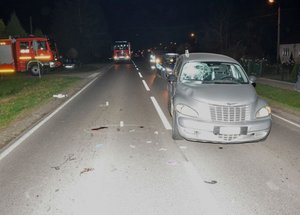 Zdjęcie przedstawiające miejsce wypadku drogowego nocą. Jezdnia, na której na pierwszym planie widoczny jest samochód koloru szarego. W tle pojazdy Straży Pożarnej oraz policyjny radiowóz.