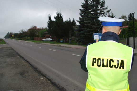 Zdjęcie kolorowe-przedstawia mały odcinek drogi i stojącego tyłem policjanta, który dokonuje pomiaru prędkości urządzeniem kontrolnym. Ubrany w mundur koloru granatowego z nałożona kamizelką odblaskową.
