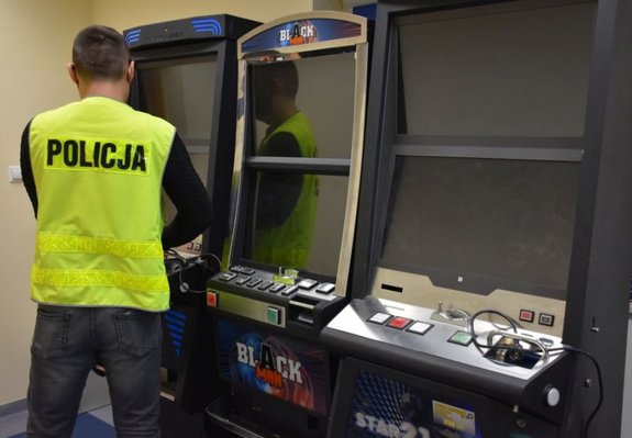 Zabezpieczone automaty do gier hazardowych. Przed nimi stoi mężczyzna w kamizelce odblaskowej z napisem Policja.
