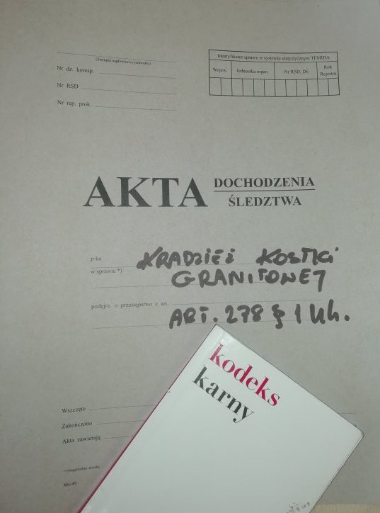 Zdjęcie przedstawia Akta dochodzeniowo – śledcze, na aktach leży Ustawa kodeks karny. Akta w szarym kolorze natomiast (Książka)Ustawa Kodeks Karny - białego koloru z napisem czerwonym Kodeks a napis Karny w czarnym