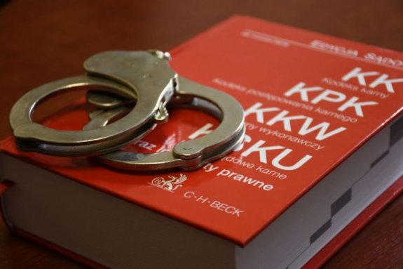 Zdjęcie kolorowe przedstawia Kodeksy KK,KPK ,KKW zawarte w jednej książce. Książka koloru czerwonego a na niej położone sa kajdanki .