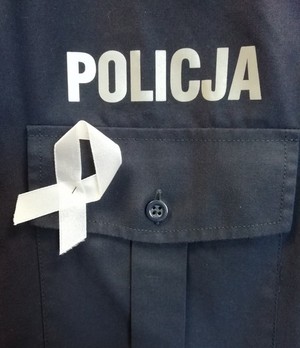 biała wstążka na granatowym tle policyjnego munduru. Powyżej biały napis policja