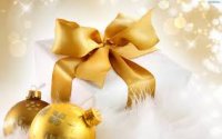 Zdjęcie kolorowe przedstawia zapakowany prezent świąteczny (pudełko owinięte w biały papier obwiązane złotą kokardą) , koło prezentu leżą dwie ozdoby choinkowe (bombki) koloru złotego.