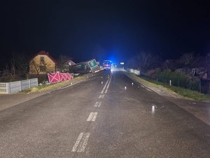 Zdjęcie w porze nocnej. Po lewej stronie drogi w rowie leżący samochód ciężarowy z naczepą, obok niego rozłożony czerwony parawan. W tle wóz strażacki.