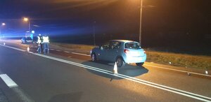 Zdjęcie w porze nocnej. Po prawej stronie drogi na pasie ruchu stoi niebieski samochód. Obok stoi dwóch policjantów w kamizelkach odblaskowych.