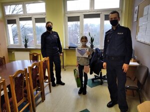 Policjant (po prawej) i policjantka (po lewej) z laureatką konkursu małą dziewczynką stojącą w środku. Dziewczynka trzyma dyplom.