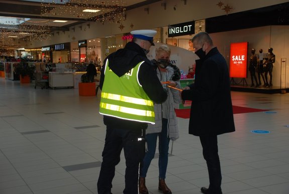 Zdjęcie przedstawia pasaż galerii Sanowej w Przemyślu . Na pierwszym planie widać policjanta ubranego w mundur oraz kamizelkę odblaskowa który wręcza opaski odblaskowe kobiecie stojącej obok niego po lewej stronie oraz mężczyźnie który stoi zaraz koło kobiety