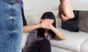Zdjęcie kolorowe przedstawia scenę z przemocy w rodzinie (kobieta siedzi przy łóżku twarz zasłania rękoma do niej podchodzi mężczyzna ma zaciśnięte pięści.