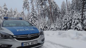 Oznakowany radiowóz policyjny, widoczny od przodu, na tle zimowej scenerii. W tle widoczne zaśnieżone drzewa iglaste.