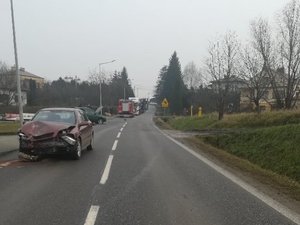 Stojący po lewej stronie pojazd marki Opel. Samochód ma uszkodzony przód. Za nim widoczny fragment pojazdu marki Renault oraz wóz strażacki.