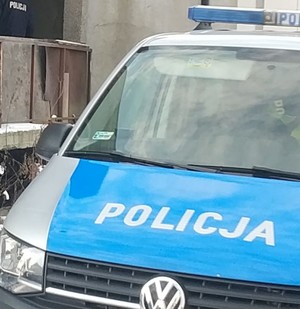 Na zdjęciu radiowóz. W tle widać budynek i policjanta.