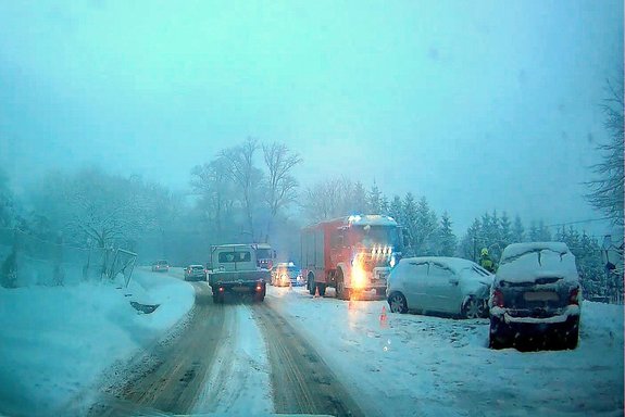Zderzenie w Wiśniowej. Na pierwszym planie dwa uszkodzone samochodu osobowe, za nimi bojowy wóz strażacki. W tle zimowa sceneria - zaśnieżona droga i drzewa.