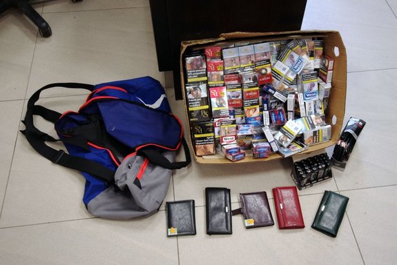 Karton z paczkami papierosów, torba turystyczna oraz portfele i zapalniczki odzyskane przez policjantów. rzeczy są ułożone na podłodze z terakoty.