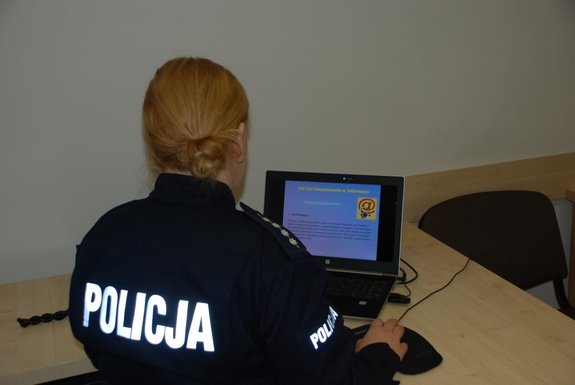 Zdjęcie kolorowe przedstawia pokój biurowy w KMP-Przemyśl przedstawia policjantkę siedzącą tyłem przy biurku na którym jest umieszczony laptop. Policjantka jest ubrana w granatowy mundur na koszulce na plecach znajduje się napis Policja.