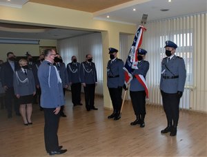 Zdjęcie przedstawia pomieszczenie w KMP-Przemyśl (aula) 
Przy ścianie okiennej stoi poczet sztandarowy (trzech policjantów ubranych ubrany w stróje galowe:czarne wysokie buty, czarne spodnie biała koszula oraz marynarka tz Gabardyna koloru siwego. Policjant stojący w środku trzyma w pozycji pion sztandar KMP-Przemyśl .Przed pocztem sztandarowym stoi Komendant Miejski Policji podinsp. Piotr Mazyr -ubrany w strój galowy:czarne buty , czarne spodnie biała koszula oraz marynarka tz Gabardyna koloru siwego.Z lewej strony widoczna Kadra kierownicza KMP-Przemyśl która ubrana jest w stroje galowe.