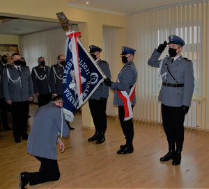 Zdjęcie przedstawia pomieszczenie w KMP-Przemyśl (aula) 
Przy ścianie okiennej stoi poczet sztandarowy (trzech policjantów ubranych ubrany w stroje galowe:czarne wysokie buty, czarne spodnie biała koszula oraz marynarka tz Gabardyna koloru siwego. Policjant stojący w środku trzyma w pozycji opuszczonej sztandar KMP-Przemyśl . Przed pocztem sztandarowym na jednym kolanie klęczy Komendant Miejski Policji podinsp. Piotr Mazyr -ubrany w strój galowy:czarne buty , czarne spodnie biała koszula oraz marynarka tz Gabardyna koloru siwego, który żegna się ze sztandarem całując go. Z lewej strony widoczna Kadra kierownicza KMP-Przemyśl która ubrana jest w stroje galowe.