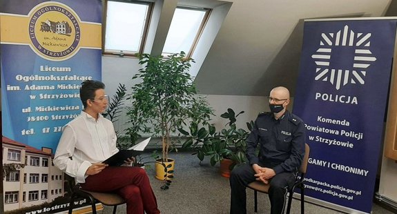 Uczeń liceum przeprowadza wywiad z policjantem. W tle banery Liceum Ogólnokształcącego w Strzyżowie i Komendy Powiatowej Policji w Strzyżowie.