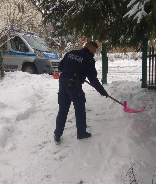 1-Zdjęcie kolorowe przedstawia posesję w miejscowości Siedliska. Na pierwszym planie widoczny policjant który odśnieża chodnik starszej kobiecie a w tle widoczny radiowóz oznakowany m-ki &quot;Dukato&quot;.