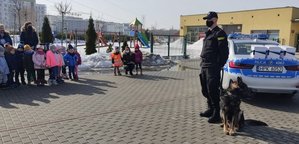 Policjant z psem służbowym stoi przed grupą dzieci. Po prawej stronie radiowóz