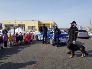 Trzej policjanci, jeden z psem służbowym, stoją przed grupą dzieci. Za funkcjonariuszami stoi radiowóz