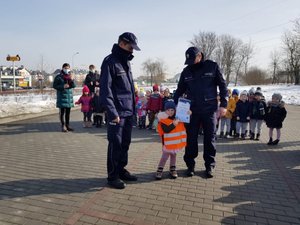Dwaj policjanci, między nimi dziewczynka z dyplomem, z tyłu grupa dzieci
