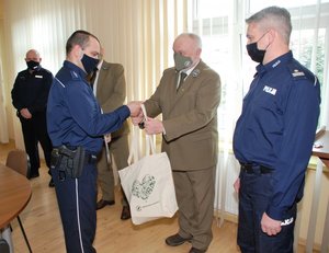 Zdjęcie przedstawia aulę w Komendzie Miejskiej Policji w Przemyślu .W tle widoczna jest okna z żaluzjami. Z prawej strony zdjęcia widoczny jest Z-ca KMP w Przemyślu Rafał Dryś koło niego stoi  Nadleśniczy Przemysław Włodek, który wręcza torbę policjantowi który stoi przed nim. Z lewej strony widoczny jest Komendant Komisariatu w Dubiecku kom. Witold Bednarczyk.