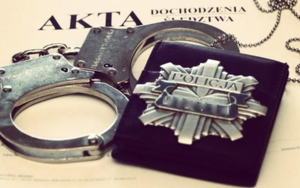 Zdjęcie kolorowe przedstawia akta policyjne na których położone są kajdanki oraz odznaka policyjna znajdująca się na czarnym etui
