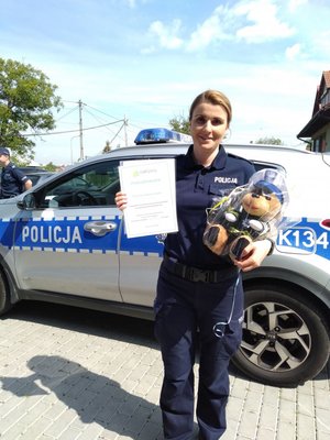 Policjantka trzyma dyplom i policyjnego misia na tle radiowozu