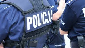 Zdjęcie kolorowe przedstawia dwóch policjantów umundurowanych z czego jeden ma na sobie kamizelkę taktyczna z napisem POLICJA. Policjanci prowadza osobę zatrzymaną Policjanci stoją tyłem