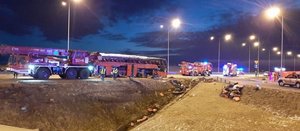 Wypadek autokaru na autostradzie A4. Pora nocna. Na pasie ruchu autostrady stoją wozy strażackie oraz strażacki dźwig. Na środku widać uszkodzony czerwony autokar i stojących obok policjantów. Na pierwszym planie rów, w którym widać leżące bagaże.