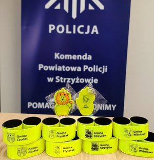 Opaski i zawieszki odblaskowe w kolorze seledynowym. w tle niebieski baner z napisem Komenda Powiatowa Policji w Strzyżowie