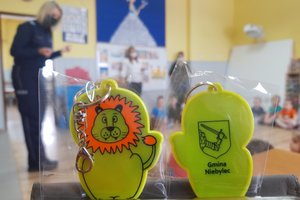 Przedszkole w Niebylcu - na pierwszym planie zawieszki odblaskowe w kształcie maskotki lwa z napisem Gmina Niebylec, w tle rozmyty obraz umundurowanej policjantki prowadzącej spotkanie z dziećmi