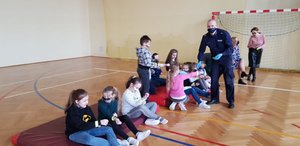 Sala gimnastyczna w Wiśniowej. umundurowany policjant wręcza dzieciom opaski odblaskowe.