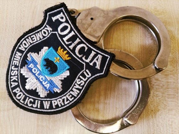 Zdjęcie kolorowe przedstawia środek przymusu bezpośredniego Kajdanki na których położony jest emblemat policyjny  z napisem U góry POLICJA a na dole widnieje napis Komenda Miejska Policji w Przemyślu. W środku widoczny niedźwiedź herb Przemyśla
