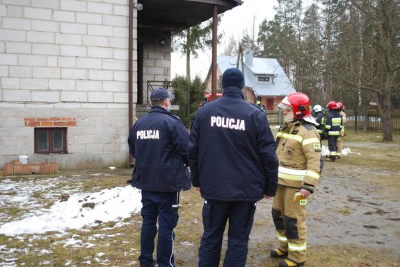 Na pierwszym planie widocznych dwóch funkcjonariuszy policji stojących obok funkcjonariuszy straży pożarnej.  W głębi budynek w którym doszło do pożaru.