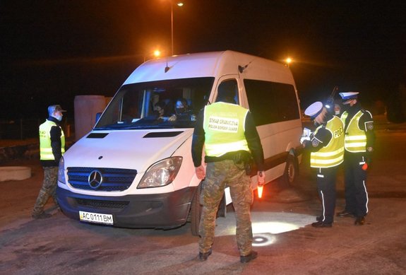 .Zdjęcie wykonane w porze nocnej -przedstawia wspólne działania Policji i Straży Granicznej . Na zdjęciu widoczny duży pojazd  w kolorze srebrnym z pasażerami w środku . Po prawej strony stoi dwóch funkcjonariuszy Policji kobieta i mężczyzna ubrani w granatowe mundury oraz na wierzchnim ubraniu mają założone kamizelki odblaskowe . Kobieta sprawdza dokumenty a mężczyzna trzyma latarkę w dłoni i umożliwia jej widoczność. Przed samochodem widoczny funkcjonariusz Straży Granicznej stojący tyłem w kamizelce odblaskowej z widocznym napisem Straż Graniczna –Polish Border Guard w prawej ręce trzyma latarkę .Z lewej strony pojazdu widoczny jest również Strażnik Graniczny ubrany w mundur moro i wierzchnia kamizelkę odblaskową. z  pora nocna .